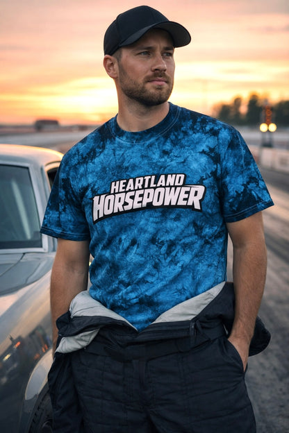 Man wearing a blue tie-dye t-shirt with 'Heartland Horsepower' text, standing next to a car at sunset.