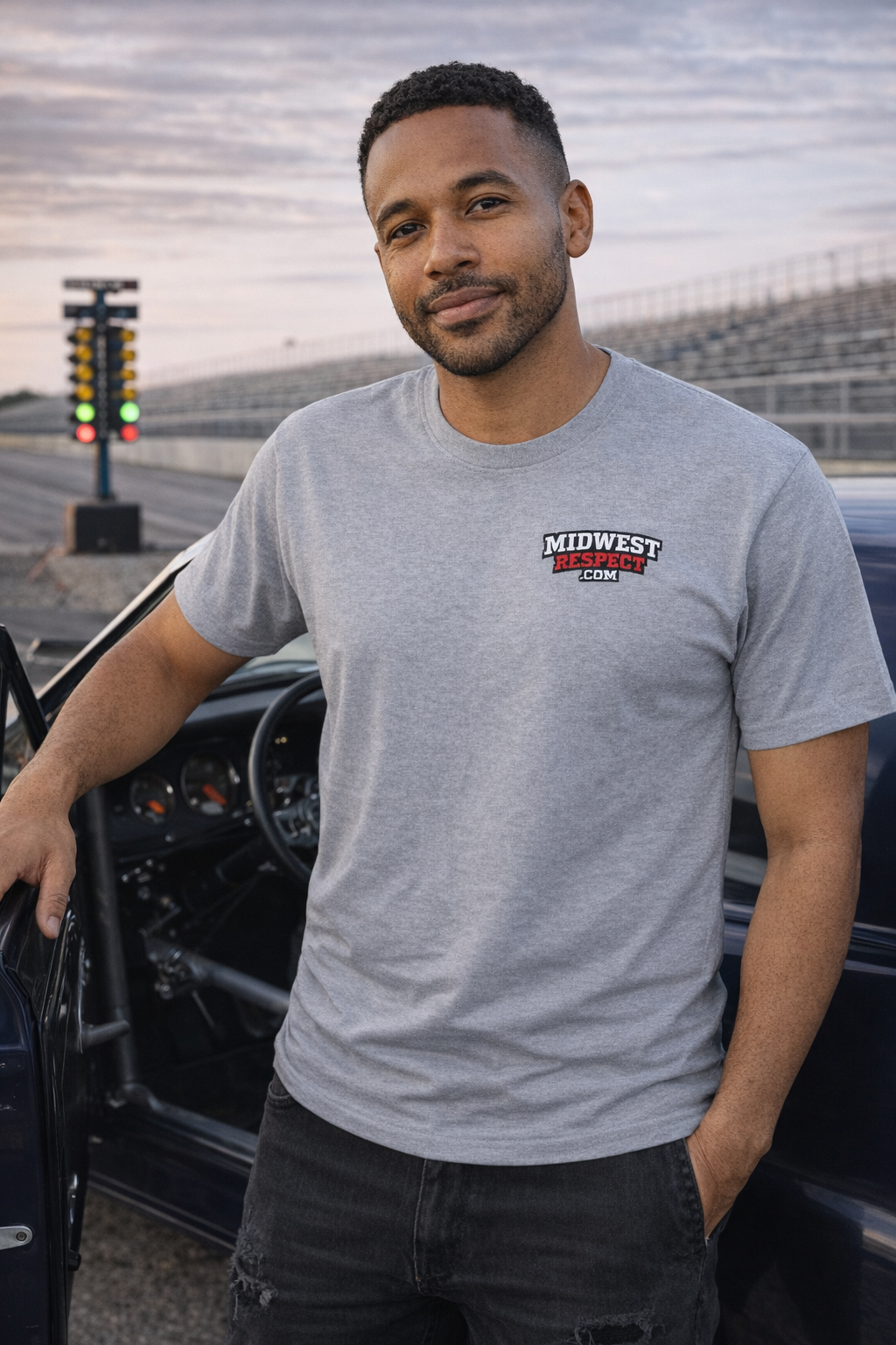 Man wearing a gray t-shirt with a logo, standing next to a car at a race track.