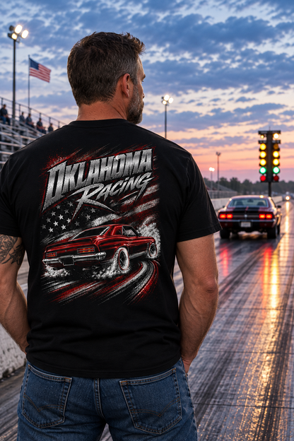 A man stands on a racetrack at dusk wearing a black graphic T-shirt with 'Oklahoma Racing' and a red muscle car print on the back, paired with blue jeans. A car is visible in the distance on the track.