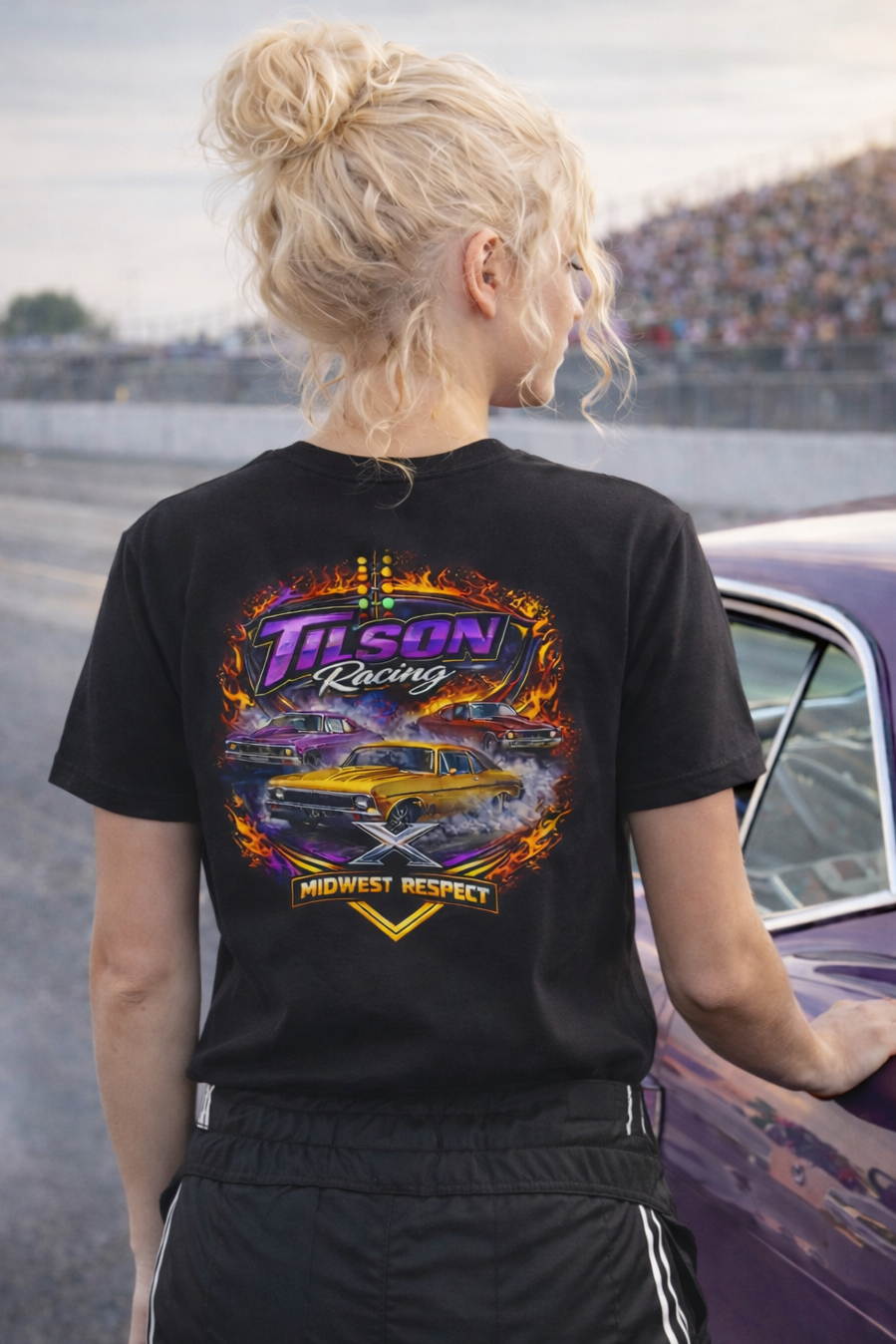 Person wearing a black t-shirt with a colorful graphic design, standing next to a purple car on a racetrack.